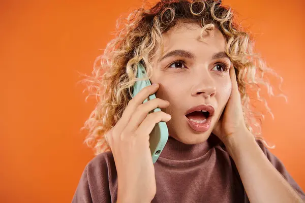 Schockierte Frau mit lockigem Haar und offenem Mund schaut weg, während sie auf dem Smartphone auf orange spricht — Stockfoto