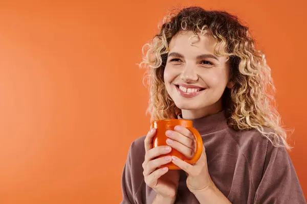 Fröhliche Frau mit lockigem Haar und einer Tasse Morgenkaffee, die auf orangefarbene, tägliche Routine blickt — Stockfoto