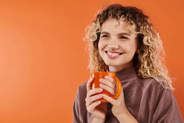 Cheerful woman with curly hair and cup of morning coffee looking away on orange, daily routine — Stock Photo