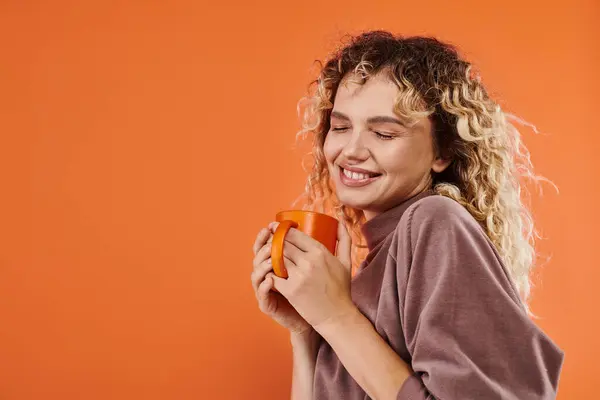 Pleased woman with curly hair and cup of morning coffee smiling with closed eyes on orange — Stock Photo