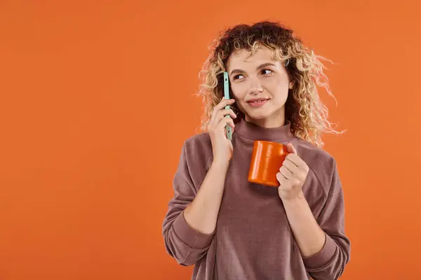 Happy curly woman with cup of morning coffee talking on smartphone on bright orange backdrop — Stock Photo