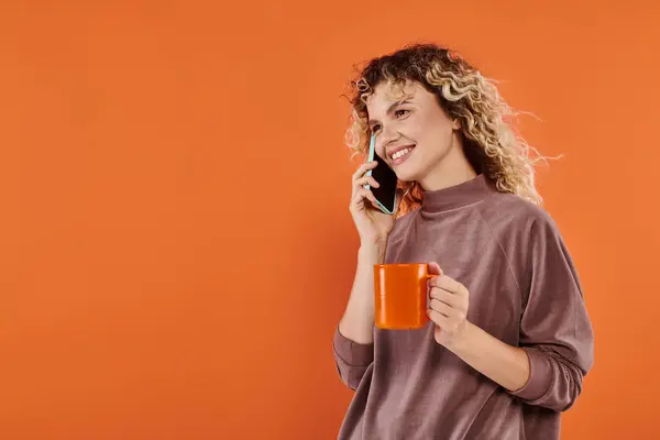 Cheerful curly woman with cup of morning coffee talking on smartphone on bright orange backdrop — Stock Photo
