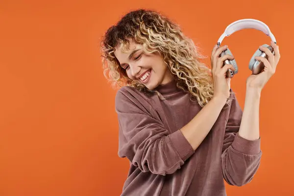 Carefree and stylish curly woman in mocha color turtleneck holding wireless headphones on orange — Stock Photo