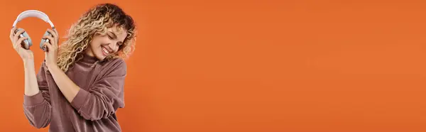Joyful and trendy woman in mocha color turtleneck holding wireless headphones on orange, banner — Stock Photo