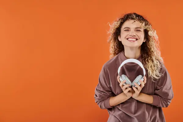 Excited curly woman in brown turtleneck posing with wireless headphones on orange backdrop — Stock Photo