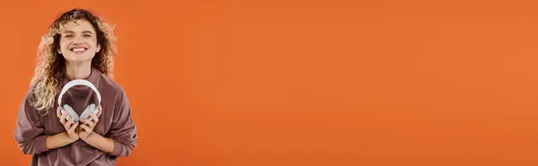 Jolly curly woman in mocha color turtleneck standing with wireless headphones on orange, banner — Stock Photo