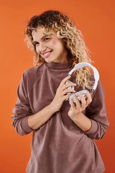 Smiling curly woman in stylish turtleneck posing with wireless headphones on orange backdrop — Stock Photo