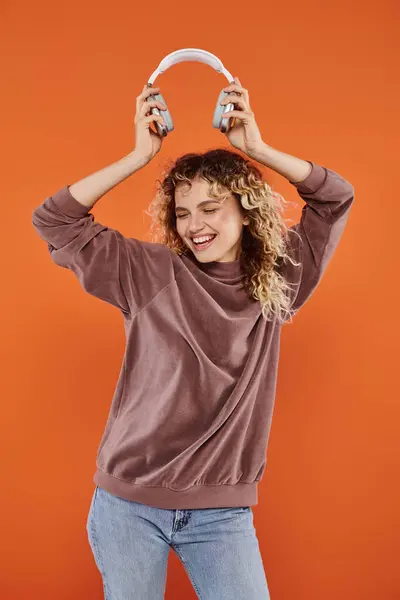 Overjoyed curly woman in stylish turtleneck dancing with wireless headphones on orange backdrop — Stock Photo