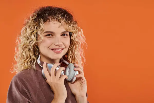 Positive curly woman in trendy turtleneck with wireless headphones looking at camera on orange — Stock Photo