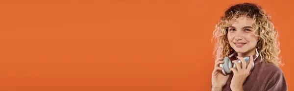 Happy curly woman in stylish turtleneck with wireless headphones looking at camera on orange, banner — Stock Photo