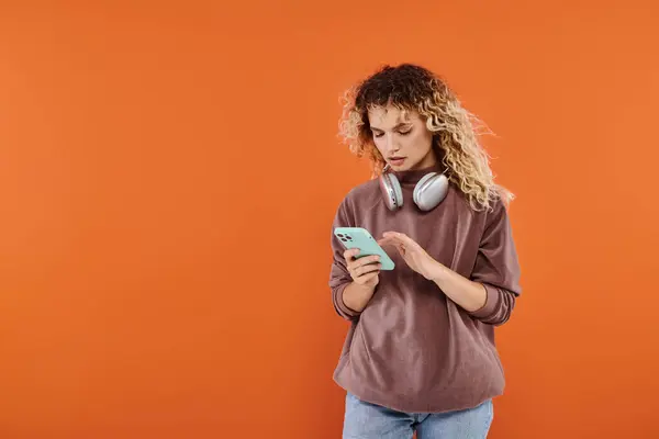 Stylish curly woman with wireless headphones messaging on mobile phone on orange backdrop — Stock Photo