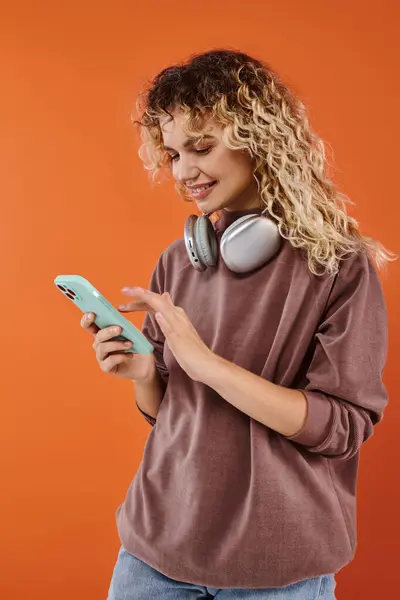 Cheerful curly woman with wireless headphones messaging on mobile phone on orange backdrop — Stock Photo