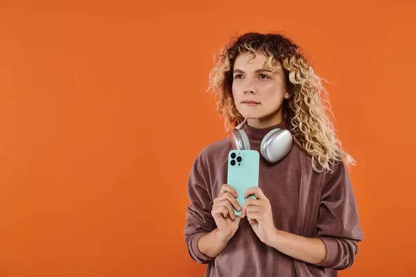 Mujer rizada reflexiva con auriculares inalámbricos y teléfono móvil mirando hacia otro lado en el fondo naranja - foto de stock