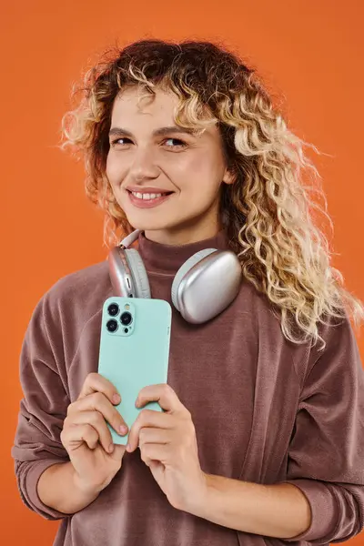 Mujer rizada alegre con auriculares inalámbricos y teléfono móvil mirando a la cámara en el fondo naranja - foto de stock