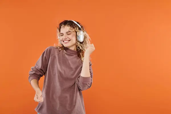 Excited woman with wavy hair dancing in wireless headphones and mocha color turtleneck on orange — Stock Photo