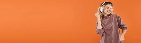 Gioiosa donna con capelli ondulati che balla in cuffia e dolcevita color moka su arancione, striscione — Foto stock