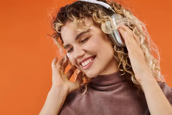 Mulher encaracolado encantado e elegante em fones de ouvido sem fio ouvir música no fundo laranja — Fotografia de Stock