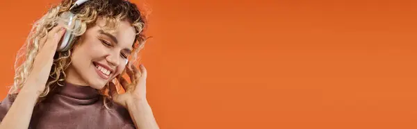 Pleased curly woman in wireless headphones listening music with closed eyes on orange, banner — Stock Photo