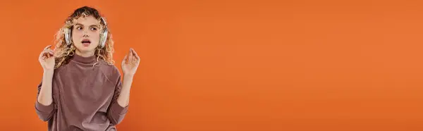 Durchdachtes Modell mit welligem Haar, das Musik in drahtlosen Kopfhörern auf orangefarbenem Hintergrund hört, Banner — Stockfoto