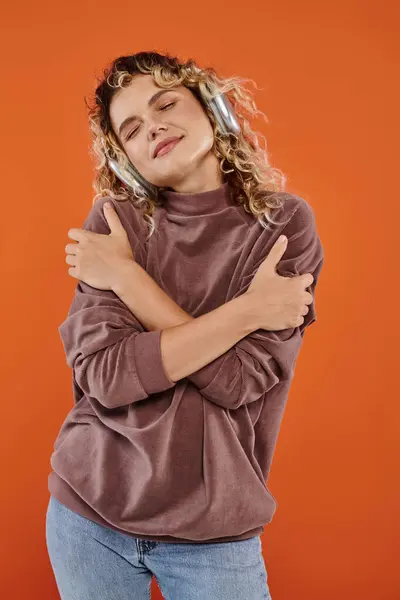 Dreamy woman with wavy hair listening music in wireless headphones and hugging herself on orange — Stock Photo