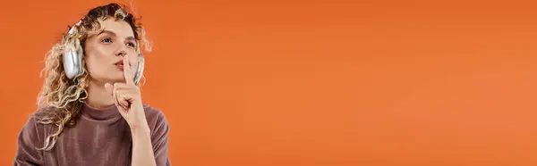 Stylische lockige Frau, die Musik in drahtlosen Kopfhörern hört und Schweigezeichen auf orangefarbenem Banner zeigt — Stockfoto