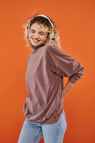 Joyful curly woman listening music in wireless headphones and smiling with closed eyes on orange — Stock Photo