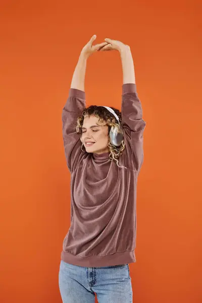 Приветливая кудрявая женщина с закрытыми глазами и поднятыми руками слушающая музыку в беспроводных наушниках на оранжевом — стоковое фото