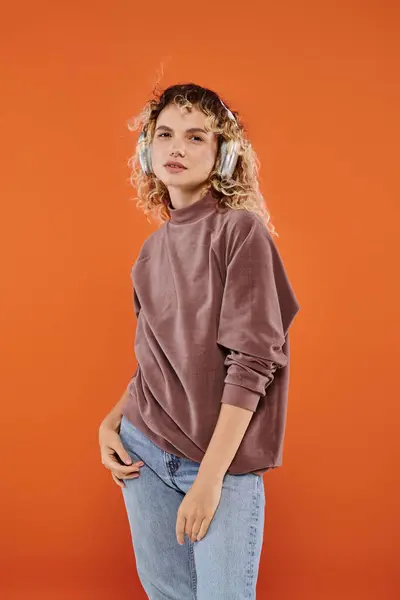Expressive woman with wavy hair in brown turtleneck listening music in wireless headphones on orange — Stock Photo