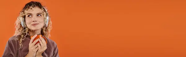 Dreamy smiling woman with cup of coffee listening music in wireless headphones on orange, banner — Stock Photo