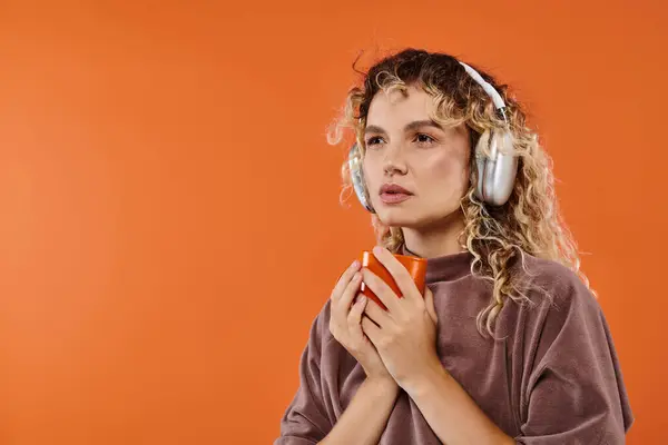 Pensive curly woman with cup of morning coffee listening music in wireless headphones on orange — Stock Photo