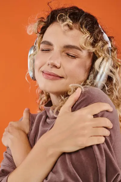 Pleased curly woman in headphones embracing herself listening music with closed eyes on orange — Stock Photo