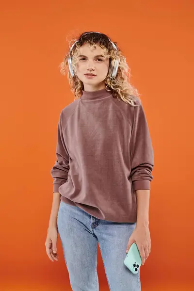 Trendy curly woman in wireless headphones holding smartphone and looking at camera on orange — Stock Photo