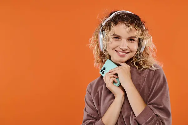 Smiling curly woman holding smartphone and listening music in wireless headphones on orange backdrop — Stock Photo