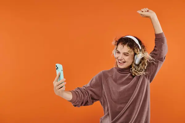 Femme bouclée joyeuse dansant dans des écouteurs sans fil et prenant selfie sur smartphone sur orange — Photo de stock