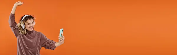 Mulher encaracolado animado dançando em fones de ouvido sem fio e tirar selfie no smartphone em laranja, banner — Fotografia de Stock