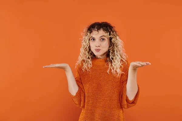 Confused curly woman in knitted sweater puffing cheeks and showing shrug gesture on orange — Stock Photo