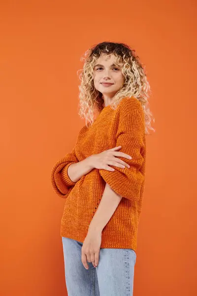 Trendfrau mit welligem Haar in kuscheligem Strickpullover blickt vor leuchtend orangefarbenem Hintergrund in die Kamera — Stockfoto