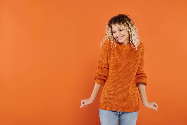 Joyous and curly woman in bright knitted sweater smiling and posing on orange studio backdrop — Stock Photo