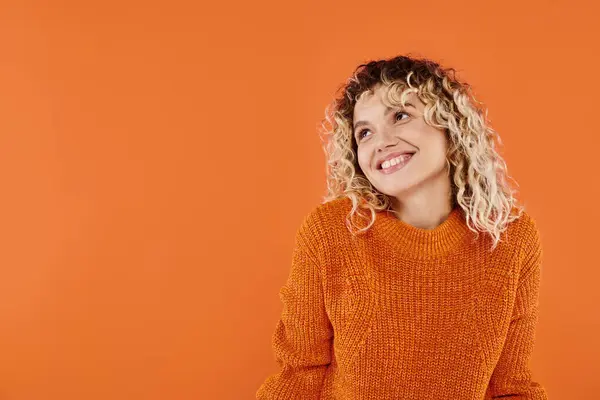 Femme heureuse et bouclée en chandail tricoté lumineux souriant regardant loin sur fond de studio orange — Photo de stock