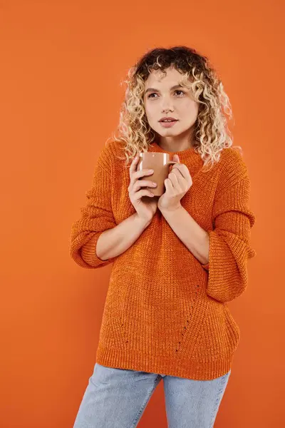 Femme réfléchie avec les cheveux bouclés et maquillage naturel tenant tasse de café sur fond orange vif — Photo de stock