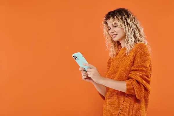Lächeln lockige Frau in hellem Strickpullover surft im Internet auf Smartphone auf orangefarbenem Hintergrund — Stockfoto