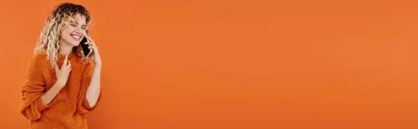 Femme frisée insouciante en chandail tricoté orange parlant sur téléphone mobile sur fond lumineux, bannière — Photo de stock