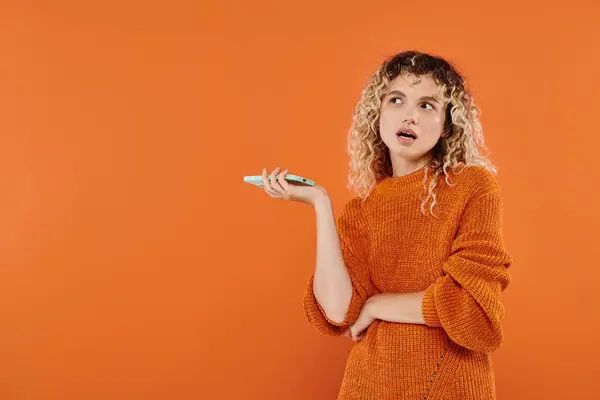 Mujer rizada reflexiva en suéter brillante sosteniendo en el teléfono móvil y mirando hacia otro lado en el fondo naranja - foto de stock