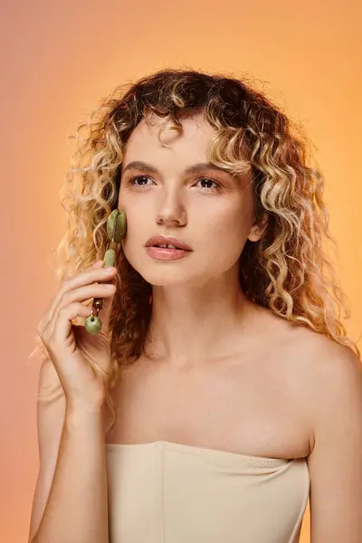 Charming woman with curly hair and perfect skin using jade roller on pink and yellow backdrop — Stock Photo