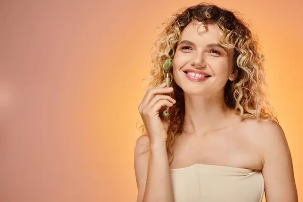 Cheerful woman with curly hair and perfect skin using jade roller on pink and yellow backdrop — Stock Photo