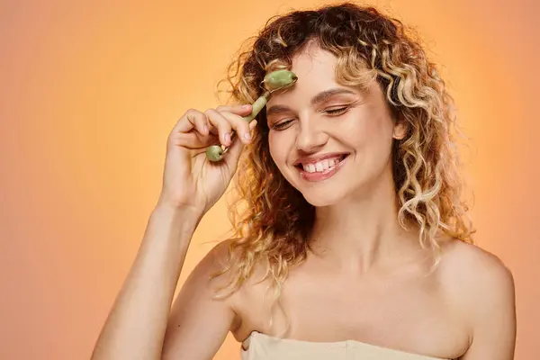 Femme ravie avec les cheveux ondulés et le visage parfait en utilisant un rouleau de jade sur fond rose et jaune — Photo de stock