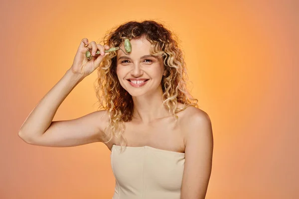 Mujer rizada feliz con la piel perfecta usando rodillo de jade y mirando a la cámara en el fondo pastel - foto de stock