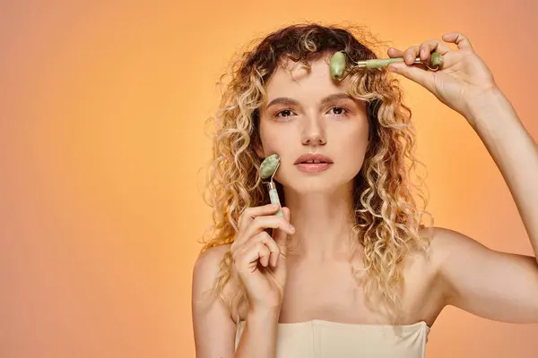 Attractive curly woman with natural makeup using jade rollers looking at camera on pastel backdrop — Stock Photo