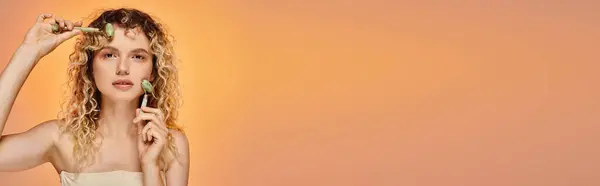 Donna riccia con pelle luminosa utilizzando rulli di giada guardando la fotocamera su sfondo pastello, banner — Foto stock