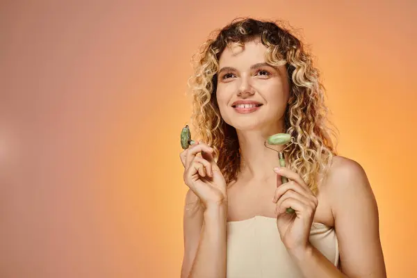 Mulher feliz e encaracolado com cabelo ondulado segurando rolos de jade e olhando para o fundo pastel — Fotografia de Stock
